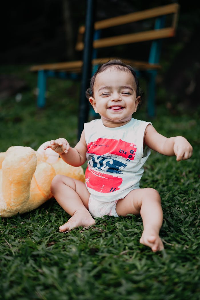 Adorable baby sitting on grass, laughing with a plush toy beside. Captures joy and innocence.